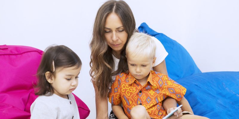 Female teacher reading a story book to two kids while sitting together in the kindergarten